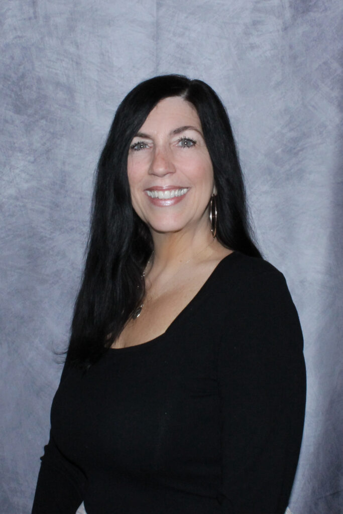 Smiling woman with long black hair wearing a black top, posing against a textured blue-gray backdrop.