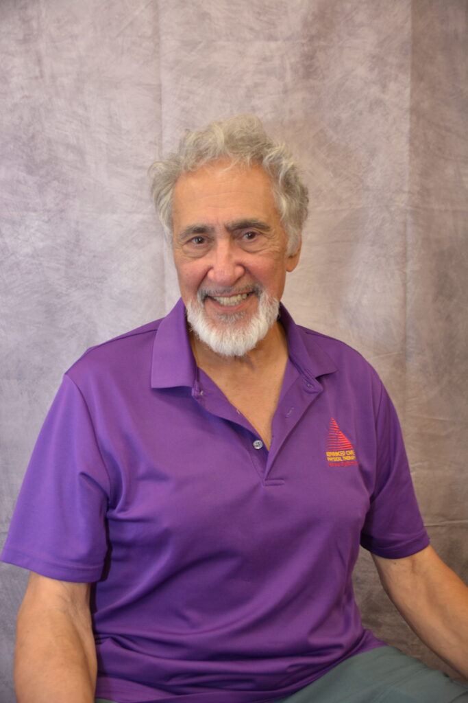 Smiling older man with gray hair and beard wearing a purple polo shirt, seated against a gray textured backdrop.