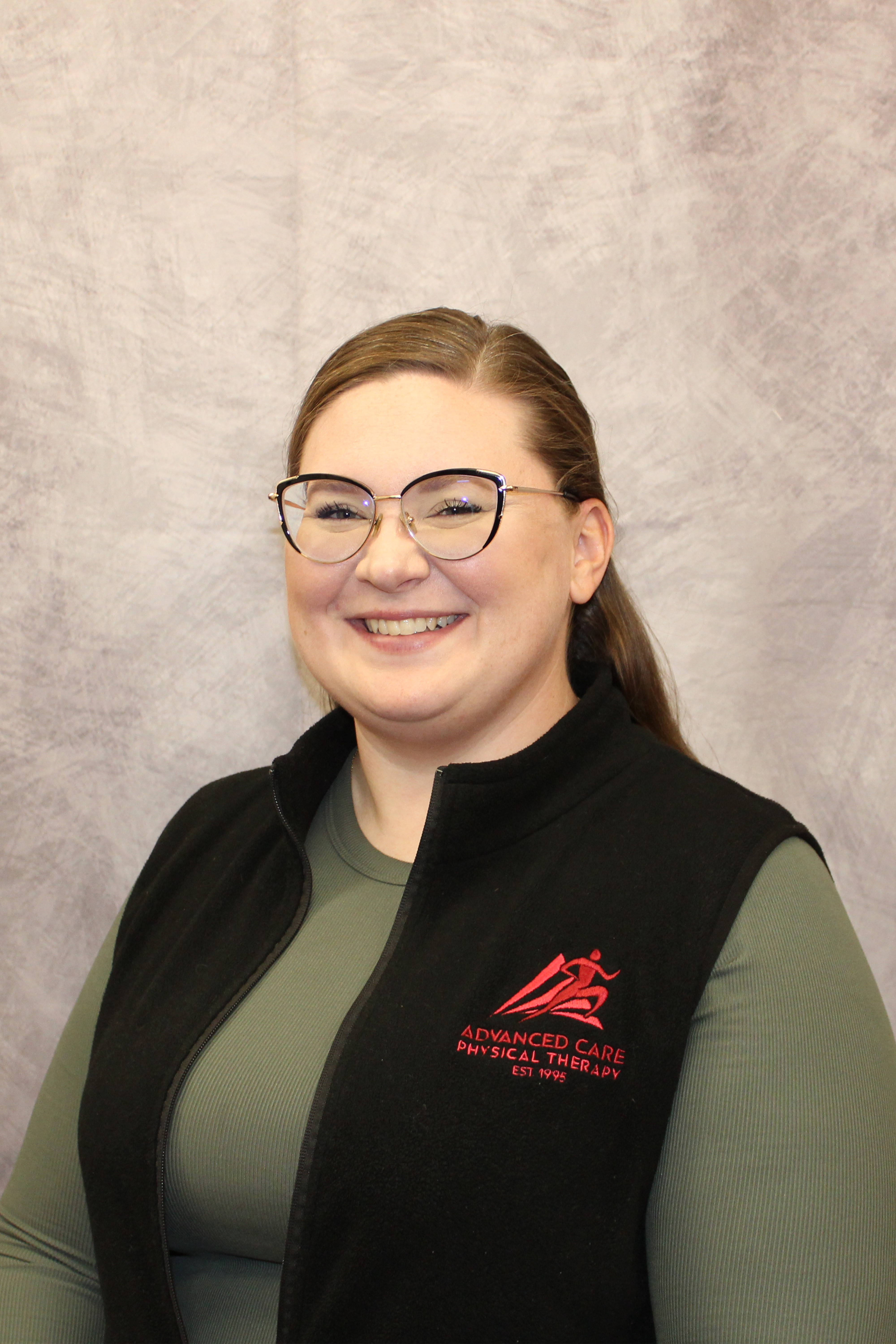 IMG_3374 Portrait - Advanced Care Physical Therapy - Advanced Care Physical Therapy Smiling woman wearing glasses and a black vest with red 'Advanced Care Physical Therapy' logo, standing against a gray textured backdrop.