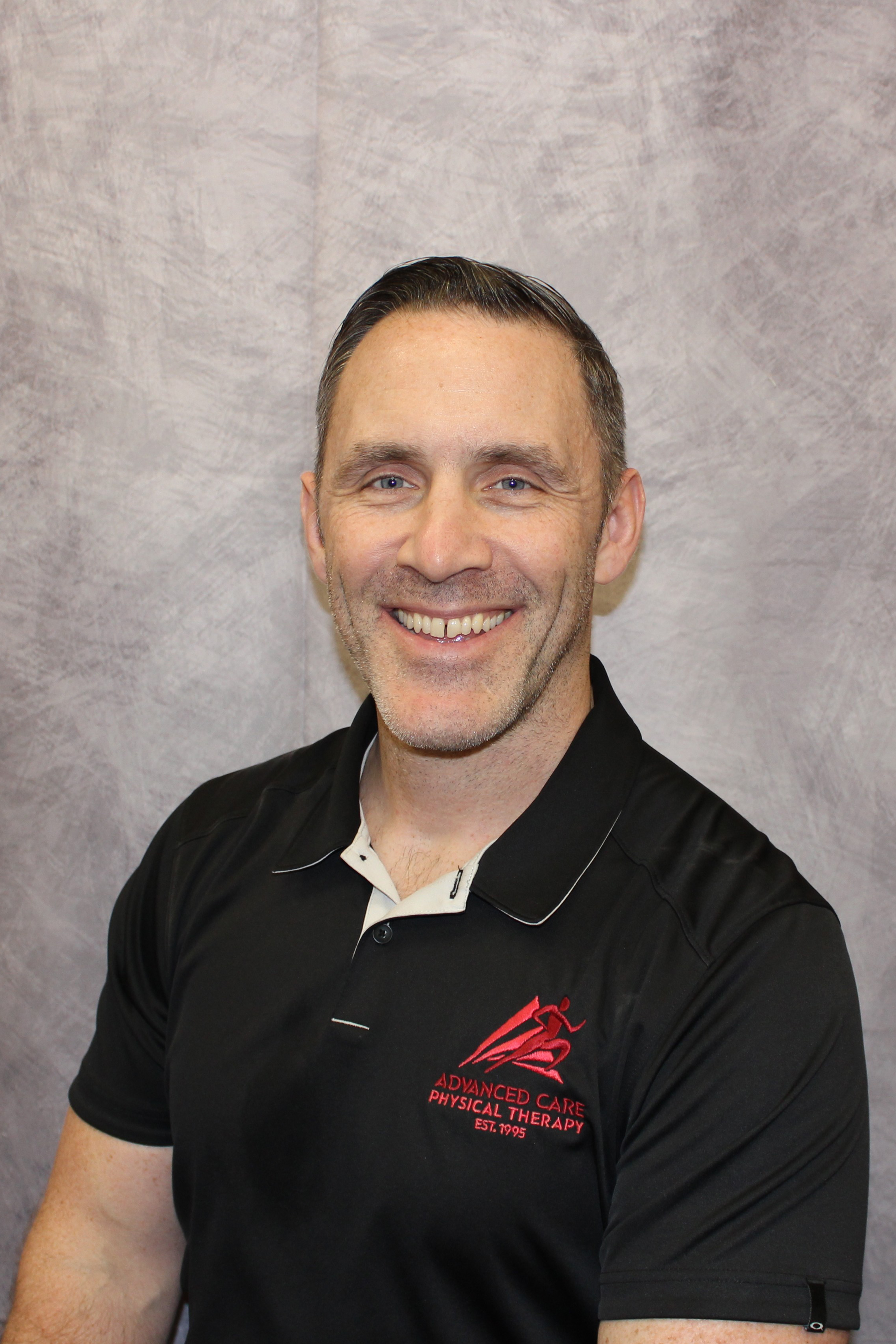 IMG_3450 Portrait - Advanced Care Physical Therapy - Advanced Care Physical Therapy Smiling man with short dark hair wearing a black polo with red logo, posed against a gray textured backdrop (therapy clinic branding).