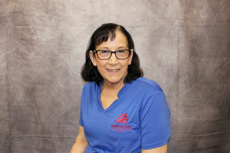 Smiling woman with black hair and glasses wearing blue scrubs against a gray backdrop.