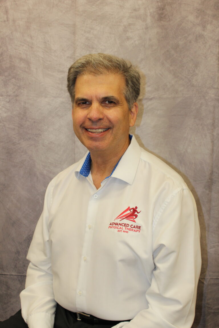 Smiling middle-aged man in a white button-down shirt with a blue inner collar, standing against a gray backdrop; logo reads 'Advanced Care Physical Therapy' on his chest poster.