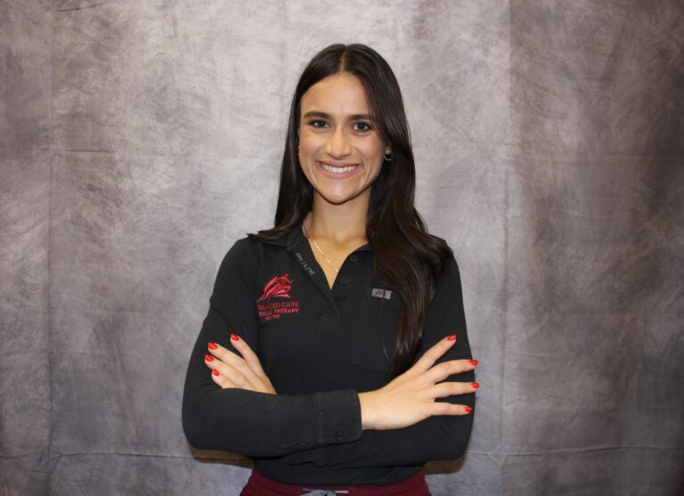 Smiling woman with arms crossed in a black polo shirt featuring a red logo, standing against a gray backdrop.