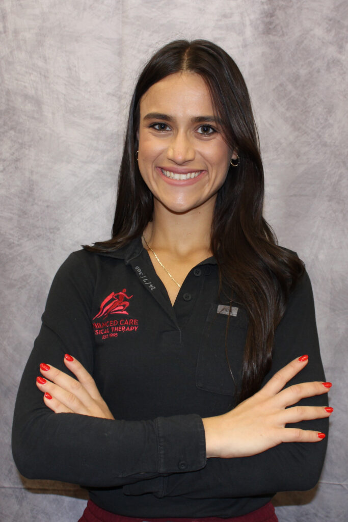 Smiling woman with long dark hair in a black polo, arms crossed, standing against a gray backdrop.