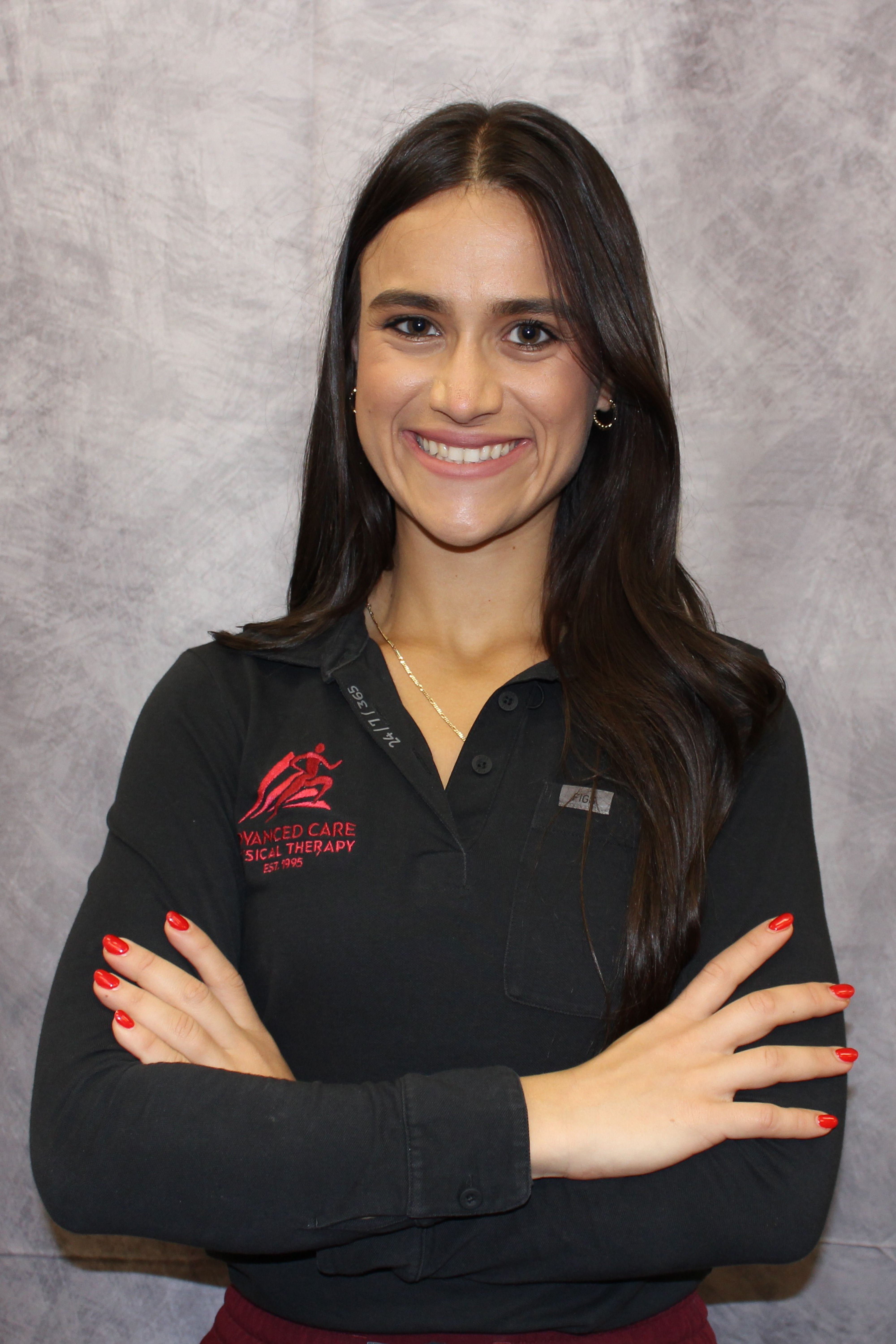 IMG_3483 Portrait - Advanced Care Physical Therapy - Advanced Care Physical Therapy Smiling woman with long dark hair in a black polo, arms crossed, standing against a gray backdrop.