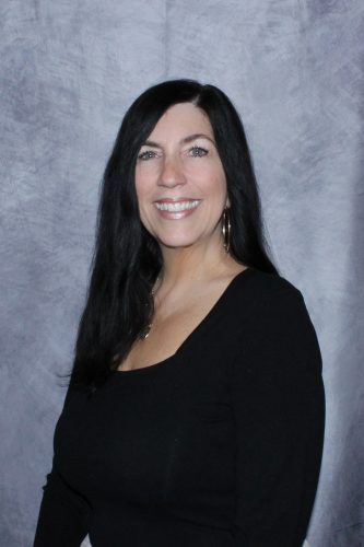Smiling woman with long black hair wearing a black top, posing against a textured blue-gray backdrop.