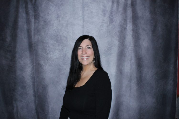 Smiling woman with long dark hair in a black top posing against a gray backdrop.