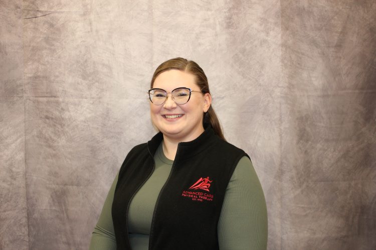 Smiling woman with glasses wearing a green shirt and black vest with a red logo that reads 'Advanced Care Physical Therapy' against a gray textured backdrop.